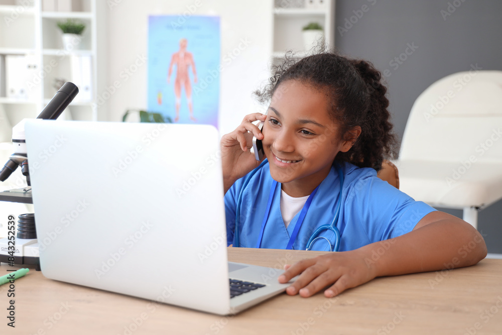 Little African-American doctor with laptop talking by mobile phone at table in clinic Little African-American doctor with laptop talking by mobile phone at table in clinic