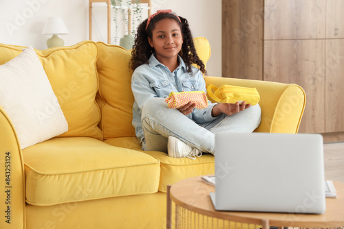 Smiling african american girl holding pencil cases on yellow sofa, home learn...