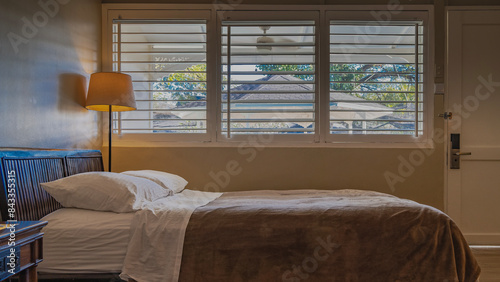 Bedroom. The bed, covered with a beige bedspread with pillows stacked at the head, stands by the window.  A floor lamp and a bedside table are nearby. The gazebo is visible through the open blinds. photo