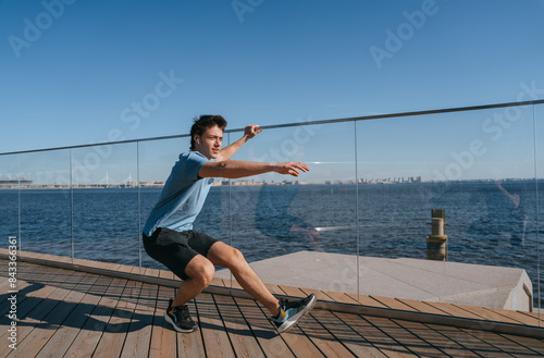 Man performing a single-leg squat outdoors by the waterfront. He is wearing a blue t-shirt and black shorts. Clear blue sky and cityscape in the background, emphasizing fitness and balance photo