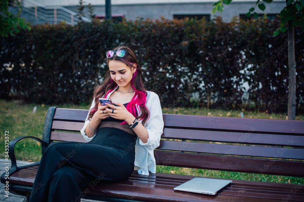 Fototapeta premium Portrait shot of a young brunette woman messaging on mobile phone while sitting on the bench outdoors. Beautiful female person using smartphone outside.