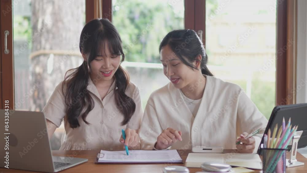 Two Asian university female friends are studying together by discussing the course content and explaining it through a laptop to help their friend understand at her home Two Asian university female friends are studying together by discussing the course content and explaining it through a laptop to help their friend understand at her home