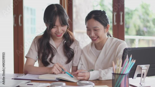 Two Asian university female friends are studying together by discussing the course content and explaining it through books and study materials to help their friend understand at her home.