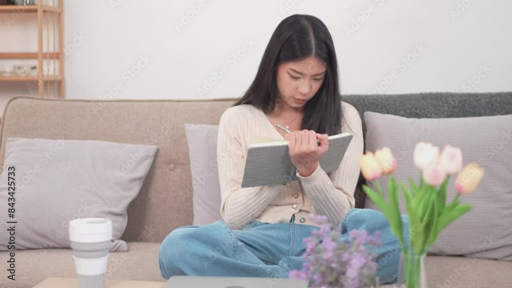 An Asian woman is writing in her diary about today's events on the sofa in her living room at home during her holiday An Asian woman is writing in her diary about today's events on the sofa in her living room at home during her holiday