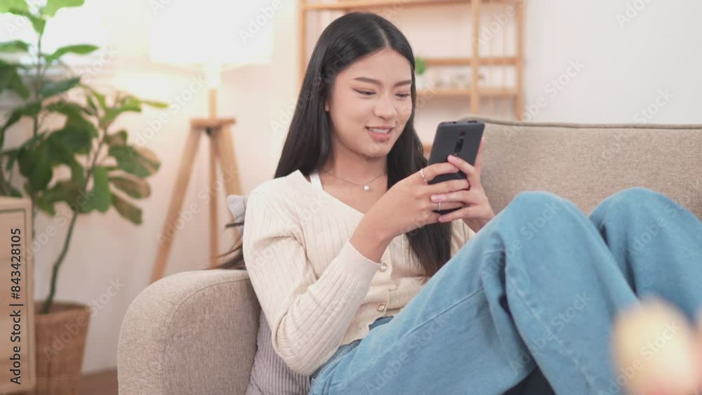 An Asian woman is lying happily on the sofa in her living room at home, playing on her smartphone through social media apps on her relaxing holiday An Asian woman is lying happily on the sofa in her living room at home, playing on her smartphone through social media apps on her relaxing holiday