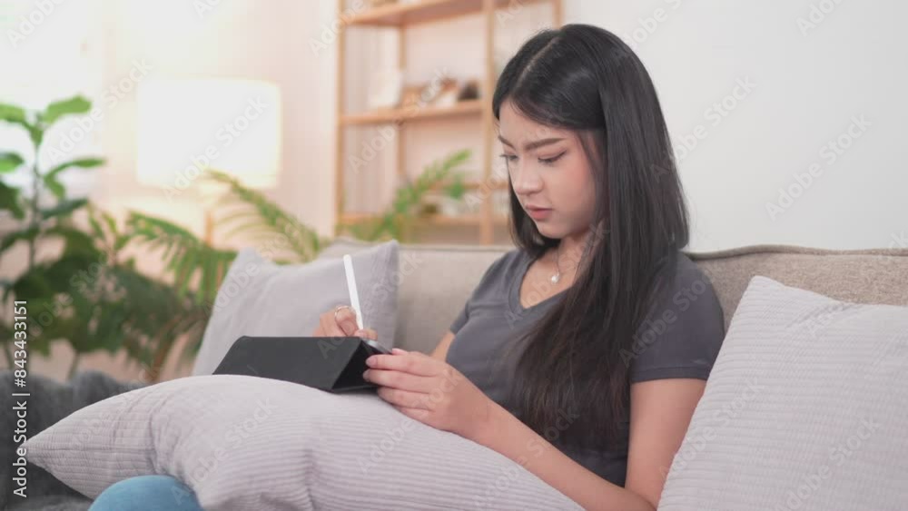An Asian female artist is happily working, drawing on her tablet, on the sofa in her living room at home during her relaxing holiday An Asian female artist is happily working, drawing on her tablet, on the sofa in her living room at home during her relaxing holiday
