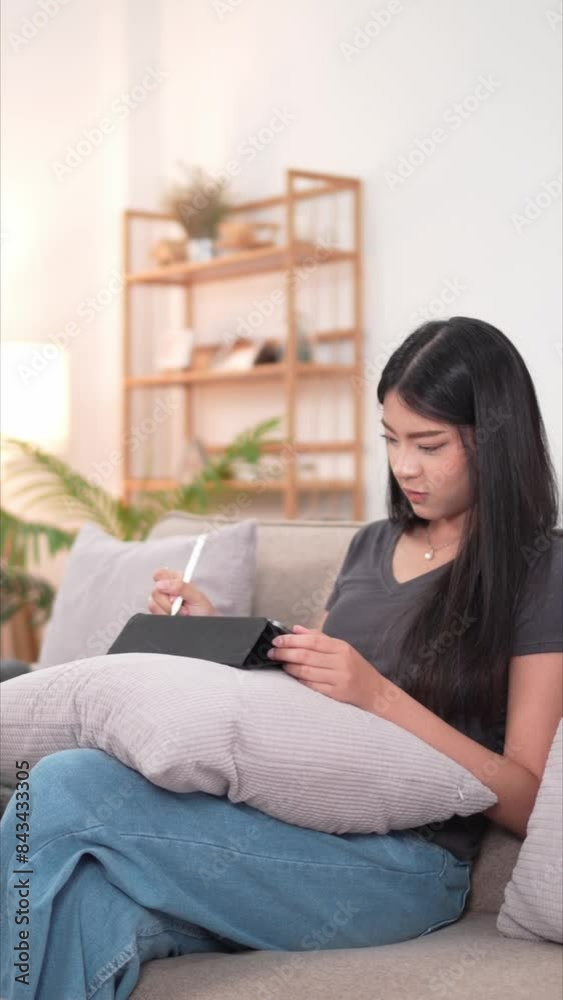 An Asian female artist is happily working, drawing on her tablet, on the sofa in her living room at home during her relaxing holiday An Asian female artist is happily working, drawing on her tablet, on the sofa in her living room at home during her relaxing holiday