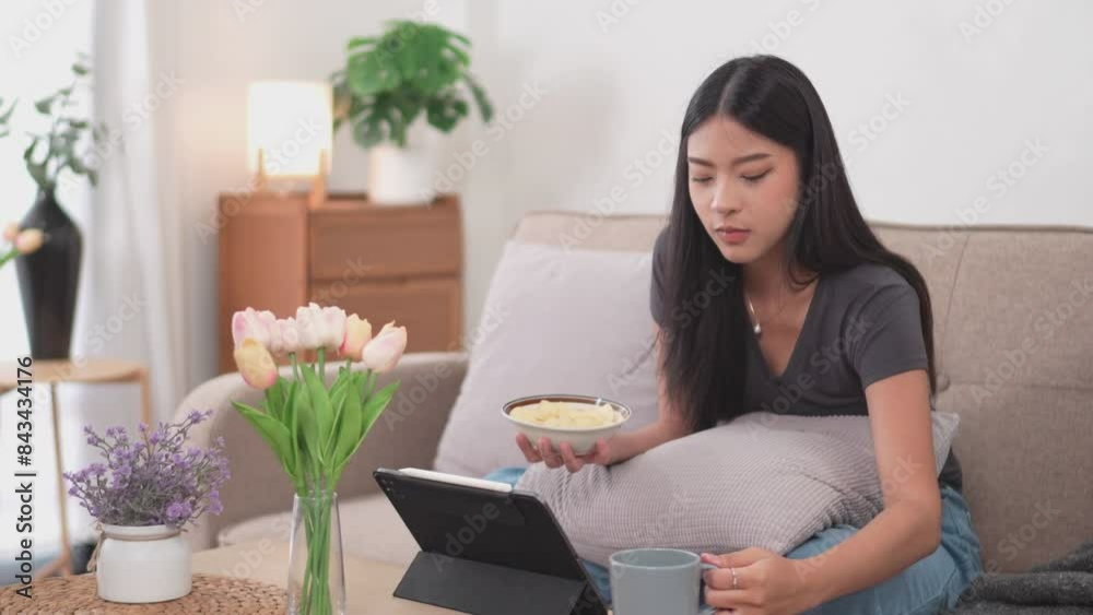 An Asian woman is happily watching a comedy series on her tablet and enjoying some snacks on the sofa in her living room at home during her relaxing holiday An Asian woman is happily watching a comedy series on her tablet and enjoying some snacks on the sofa in her living room at home during her relaxing holiday