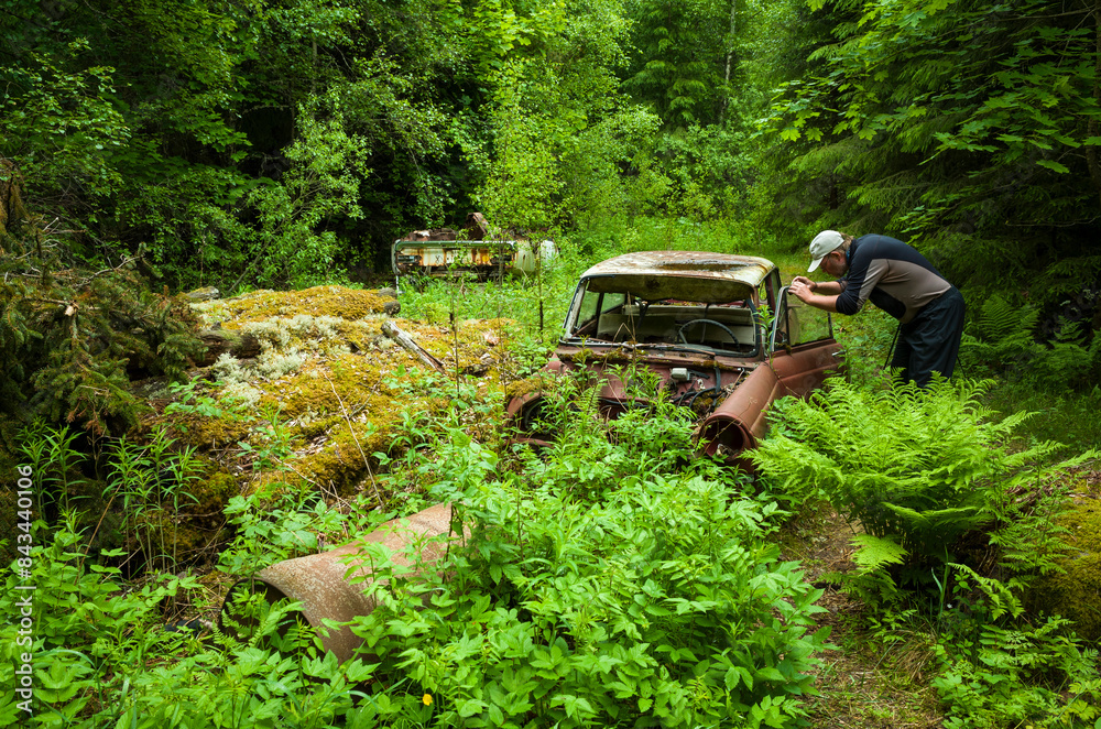 Scrap yard in forest, Man photographing an old abandoned car overgrown with lush green vegetation, Båstnäs Car Cemetery in Sweden Scrap yard in forest, Man photographing an old abandoned car overgrown with lush green vegetation, Båstnäs Car Cemetery in Sweden