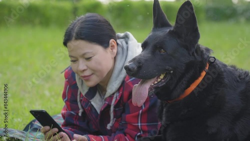 Side view of attractive mature Asian woman networking online on mobile phone, petting lovely east european black female shepherd while enjoying leisure together with dog in summer nature.