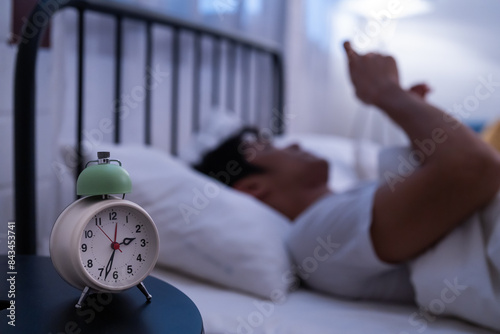 Asian man lying in bed while using his phone for surf the internet and see feed on social media when notification alert message with an alarm clock on the bedside table bedtime late night