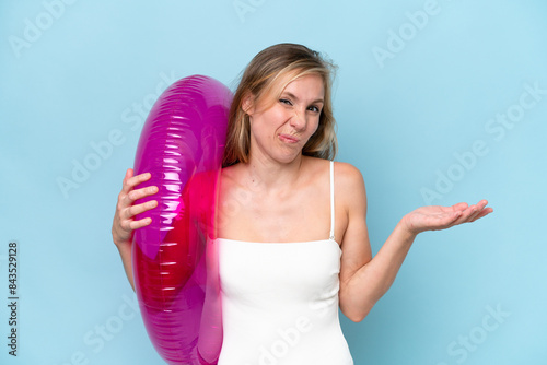 Young blonde woman in swimsuit holding inflatable donut isolated on blue background making doubts gesture while lifting the shoulders