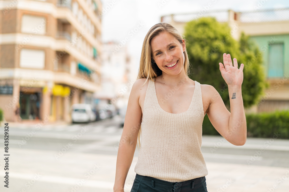 Young blonde woman at outdoors saluting with hand with happy expression