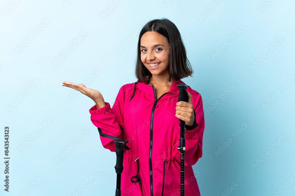 Young latin woman with backpack and trekking poles isolated on blue background extending hands to the side for inviting to come Young latin woman with backpack and trekking poles isolated on blue background extending hands to the side for inviting to come