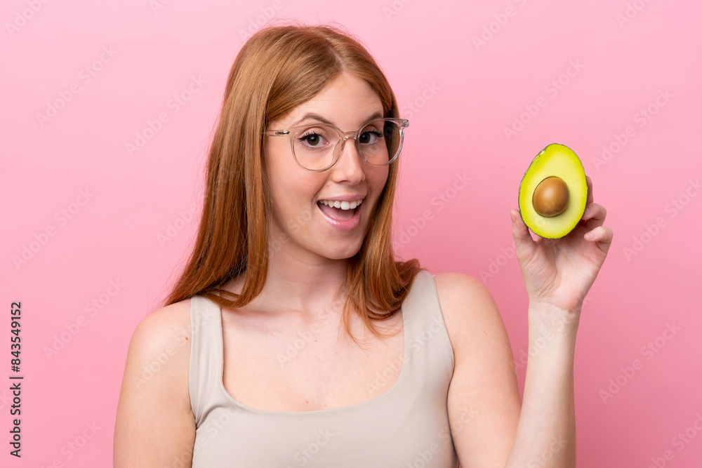 Young redhead woman holding an avocado isolated on pink background with surprise facial expression Young redhead woman holding an avocado isolated on pink background with surprise facial expression