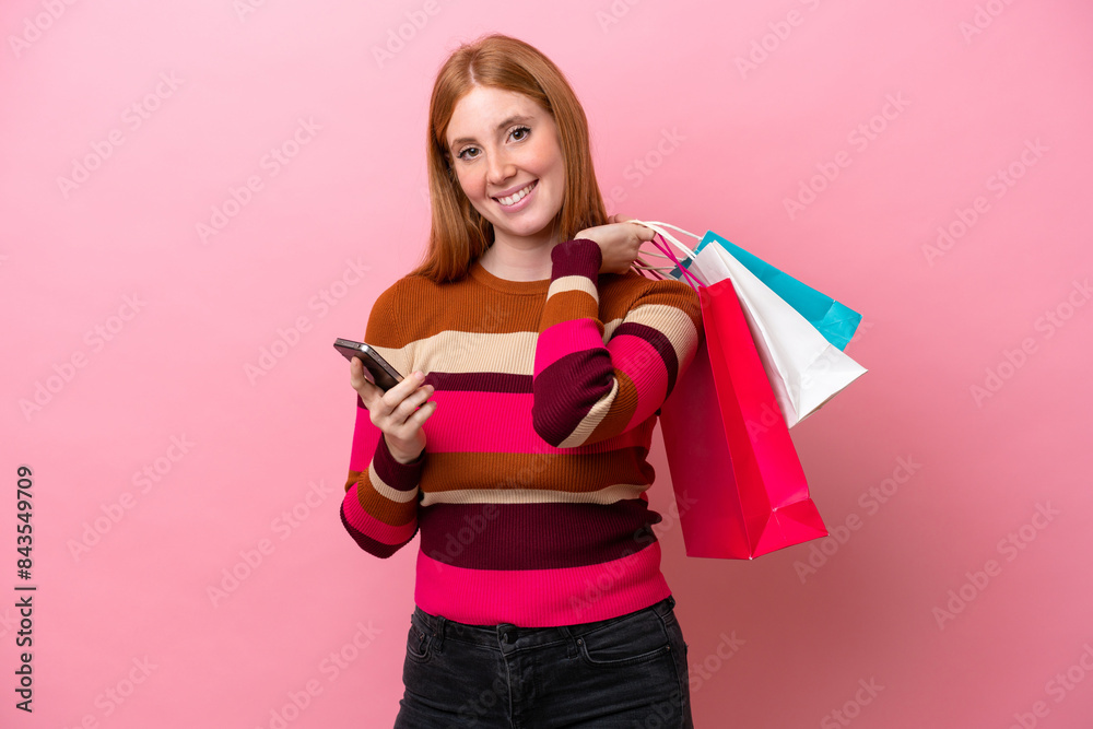 Young redhead woman isolated on pink background holding shopping bags and writing a message with her cell phone to a friend Young redhead woman isolated on pink background holding shopping bags and writing a message with her cell phone to a friend