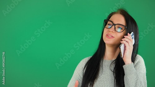 A young, beautiful girl in glasses, with black hair, of European appearance, talks on the phone, smiles and gesticulates emotionally. Green background. photo