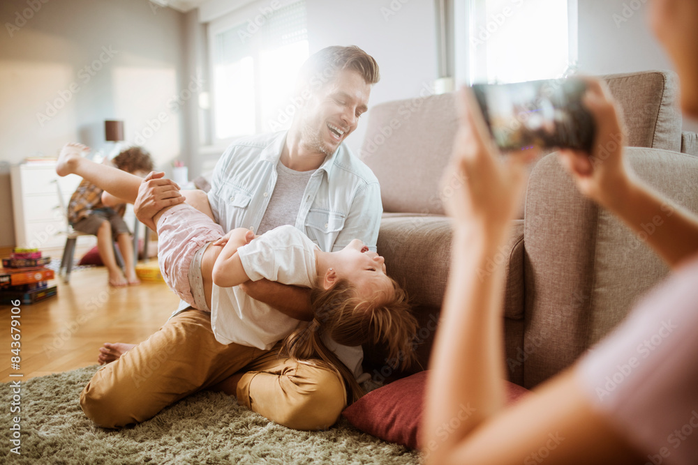 Joyful father playing with daughter while mother takes photo with smartphone at home Joyful father playing with daughter while mother takes photo with smartphone at home