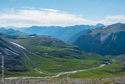 Foto Aerial top view to sunlit green alpine valley with creek among big hills and large rocky mountains silhouettes in bright sun under clouds in blue sky