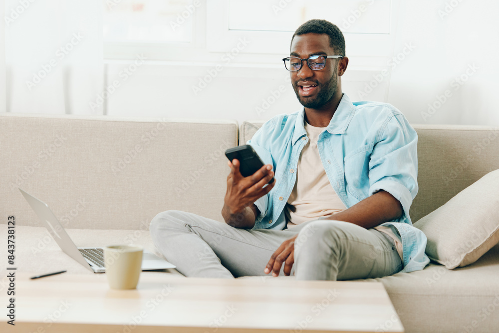 Smiling African American man working on a laptop while sitting on a sofa in his modern apartment He is wearing glasses and typing on the computer, immersed in online communication and online shopping
