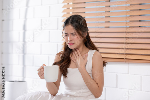 Asian woman in a white tank top holding a cup, showing discomfort and touching her throat, Health care concept photo