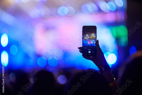 фотографія Concertgoer films live music event with smartphone, hand raised above crowd, capturing stage lights, performers in blurred background