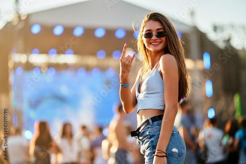 Joyful young woman enjoys summer music festival on beach, waves hand sign with stage lights, crowd in background. Trendy outfit, sunglasses, vibrant atmosphere at coastal event for promo. photo