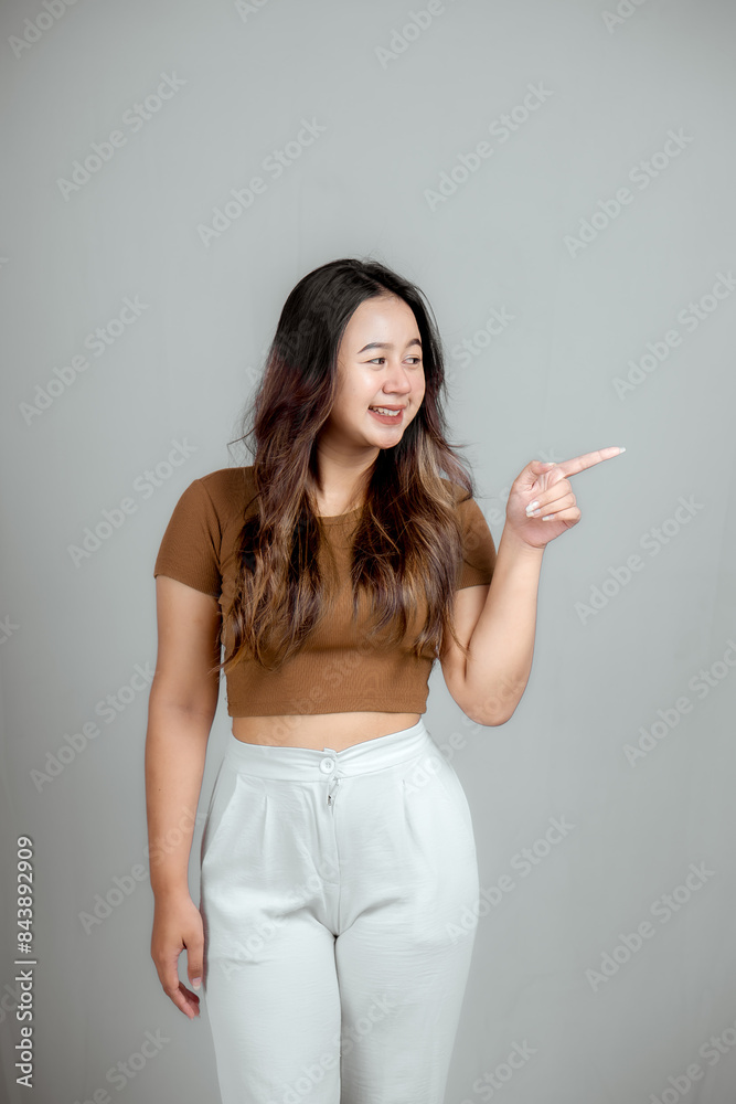 Young asian woman in surprise looks to the side and shows with index finger. Attention concept.
