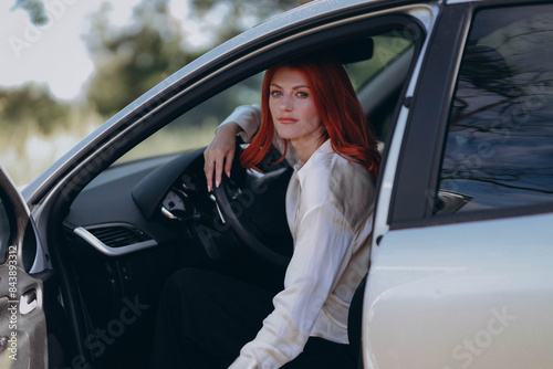 red-haired woman sits confidently in the driver's seat of a grey sedan, her face framed by cascading curls. The car's interior reflects soft daylight, highlighting her thoughtful expression in this po © Евгения Жигалкина