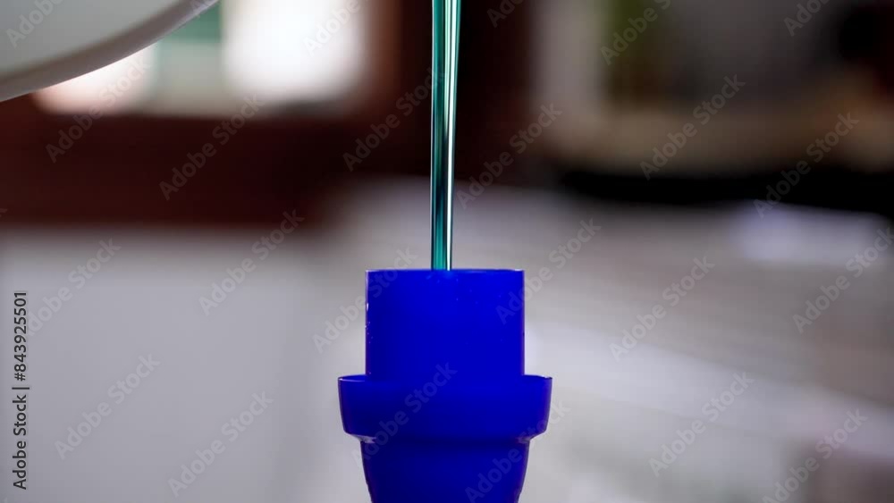 Pouring liquid blue detergent into a measuring cap for a washing machine in a room with light shining from a window. Routine household chores action