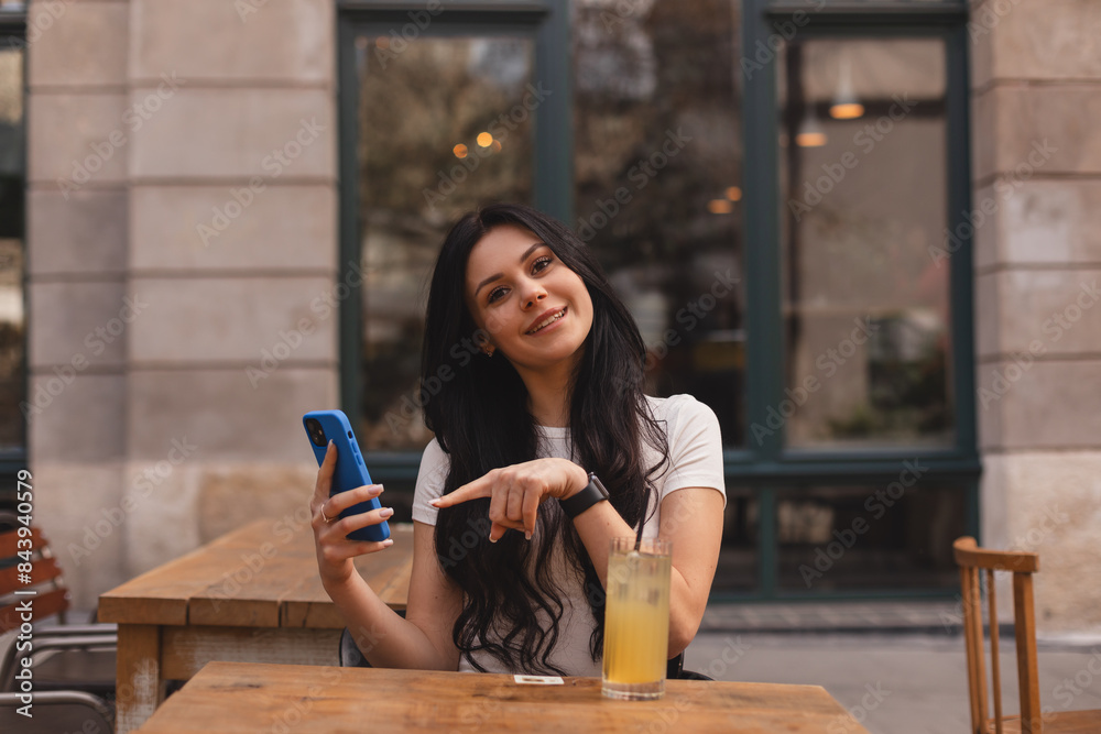 Attractive brunette woman sitting in cafe outdoors, drink cocktail, holding mobile phones, pointing index finger on screen telephone, girl wear white t-shirt. Happy smiling girl. Attractive brunette woman sitting in cafe outdoors, drink cocktail, holding mobile phones, pointing index finger on screen telephone, girl wear white t-shirt. Happy smiling girl.