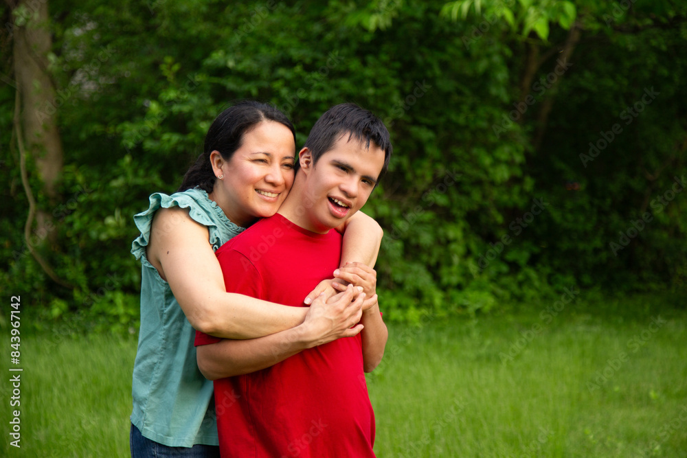 A mother and son with down syndrome embrace in a park. The woman is wearing a green T-shirt and the young man is wearing a red T-shirt. Both are smiling. A mother and son with down syndrome embrace in a park. The woman is wearing a green T-shirt and the young man is wearing a red T-shirt. Both are smiling.