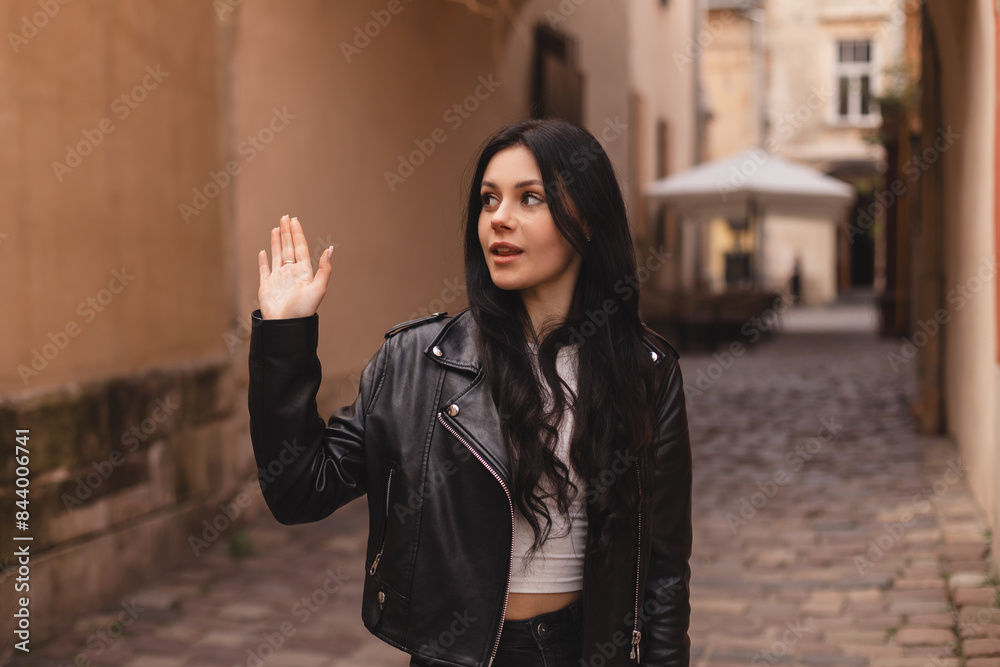 Fototapeta premium Happy attractive woman in black leather jacket, walk outdoors, meeting her friends or love, showing high five hey hello hi sign. She is joyful to meet her acquainted by chance. Girl wave her hand.