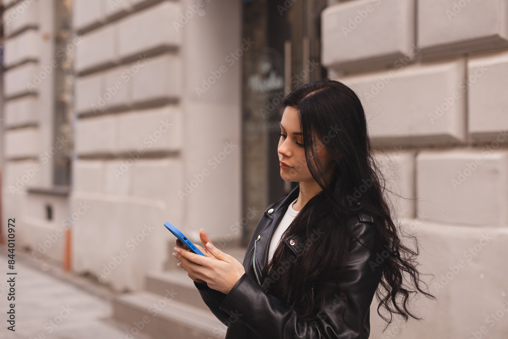 Fototapeta premium Outdoor shot of serious woman uses modern gadgets walks at street uses mobile phone, wear leather jacket sends sms tries to find route in unknown place in the city. Girl hold mobile phone and chatting