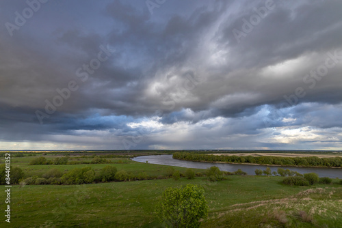 A panoramic view of a meandering river under a dramatic, stormy sky, with lush greenery and grasslands along its banks.