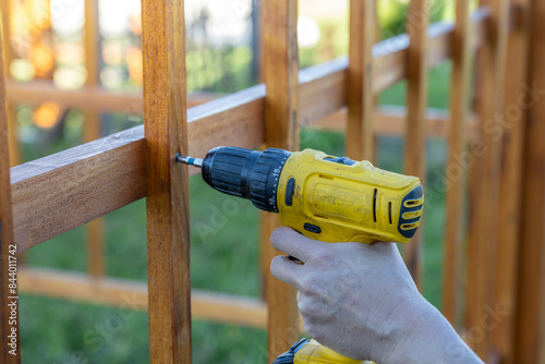 A person is using a drill to attach a wooden fence. The individual is focused on securing the fence panels to the posts using screws