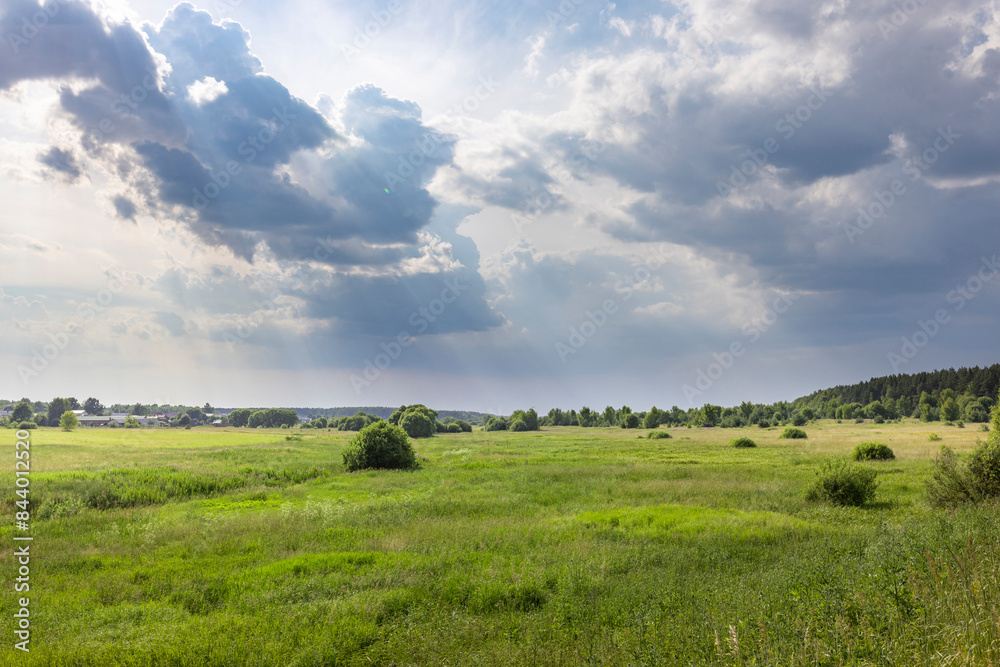 A wide shot of a green meadow with a few trees scattered throughout. The sky is dominated by large, dark clouds with patches of sunlight breaking through. A wide shot of a green meadow with a few trees scattered throughout. The sky is dominated by large, dark clouds with patches of sunlight breaking through.