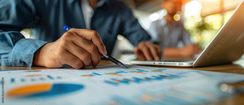 A person analyzing business charts and graphs on paper with a pen, while using a laptop, in a modern office setting with natural light. A person analyzing business charts and graphs on paper with a pen, while using a laptop, in a modern office setting with natural light.