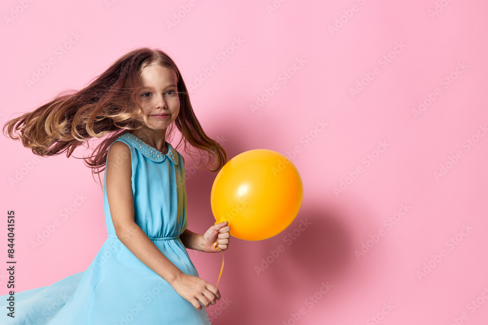 Little girl in blue dress holding orange balloon against vibrant pink background Little girl in blue dress holding orange balloon against vibrant pink background