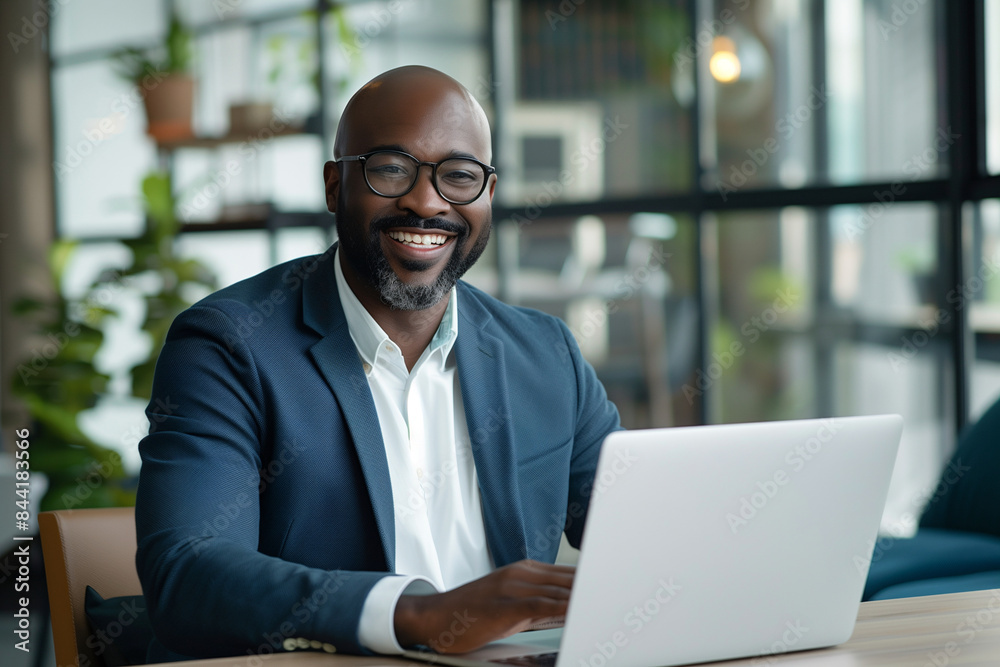 Black man in a suit is smiling and sitting at a table with a laptop. He is happy and content while working on his computer Black man in a suit is smiling and sitting at a table with a laptop. He is happy and content while working on his computer