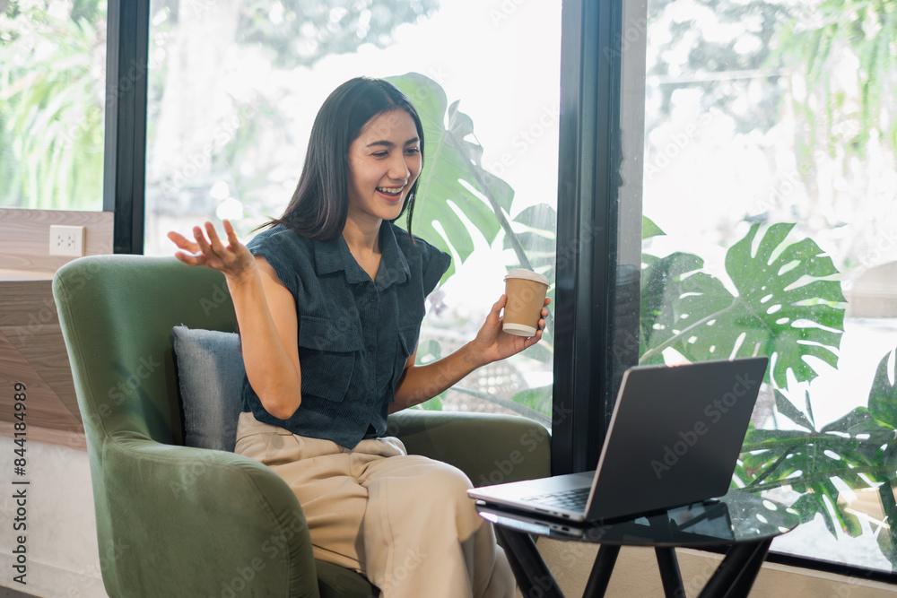 Naklejka premium A woman is sitting in a chair with a laptop open in front of her. She is holding a cup of coffee and she is smiling. The scene suggests a casual and relaxed atmosphere