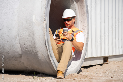 A man in a hard hat is sitting in a hole with his cell phone wearing an orange vest. A construction worker in helmet and vest sits with a coffee and a phone on his lunch hour near a construction site