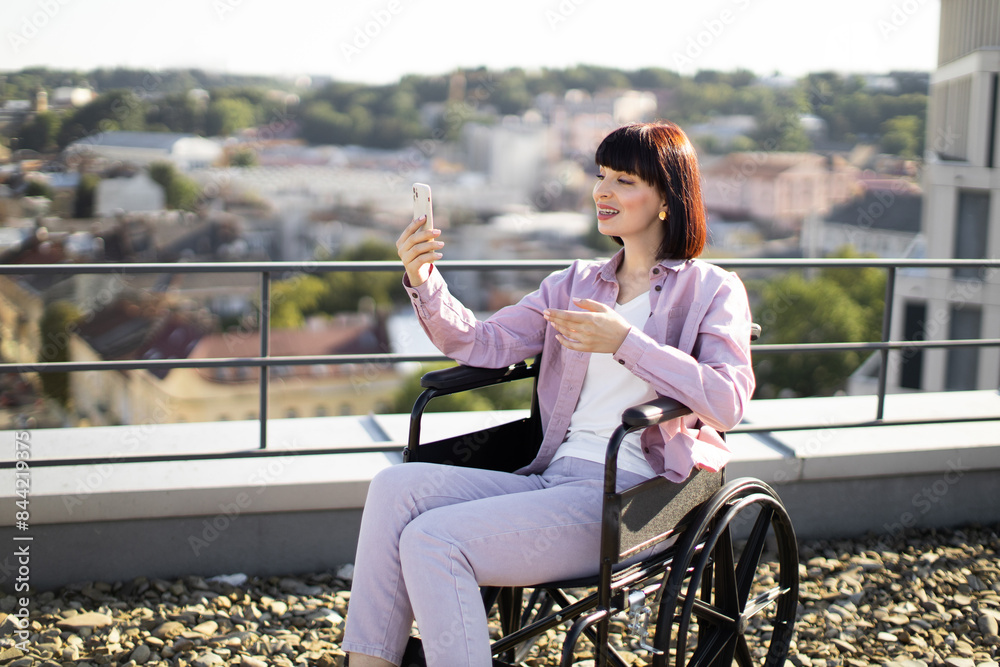 Young white woman in wheelchair enjoying video chat on smartphone while sitting on rooftop terrace overlooking city. She appears happy and content, with a beautiful urban landscape in the background. Young white woman in wheelchair enjoying video chat on smartphone while sitting on rooftop terrace overlooking city. She appears happy and content, with a beautiful urban landscape in the background.