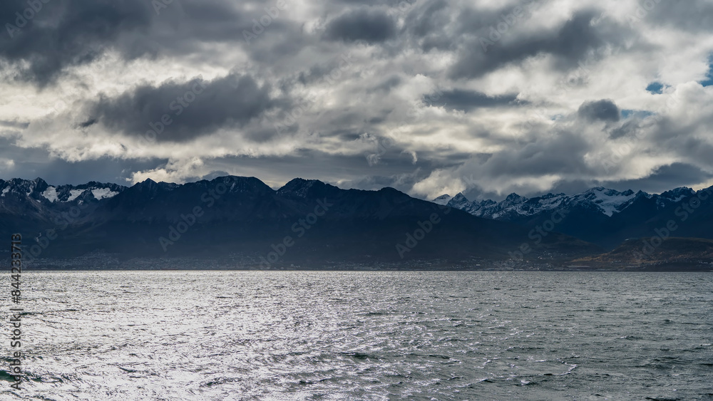 The snow-covered mountain range of the Andes Martial against the sky and clouds. The houses of Ushuaia at the foot. In the foreground is the Beagle Channel. Ripples on the shiny surface of the water. The snow-covered mountain range of the Andes Martial against the sky and clouds. The houses of Ushuaia at the foot. In the foreground is the Beagle Channel. Ripples on the shiny surface of the water.