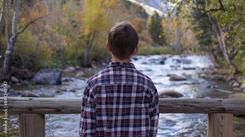 A boy wearing a plaid shirt stands facing away on a wooden bridge over a river gazing at the water