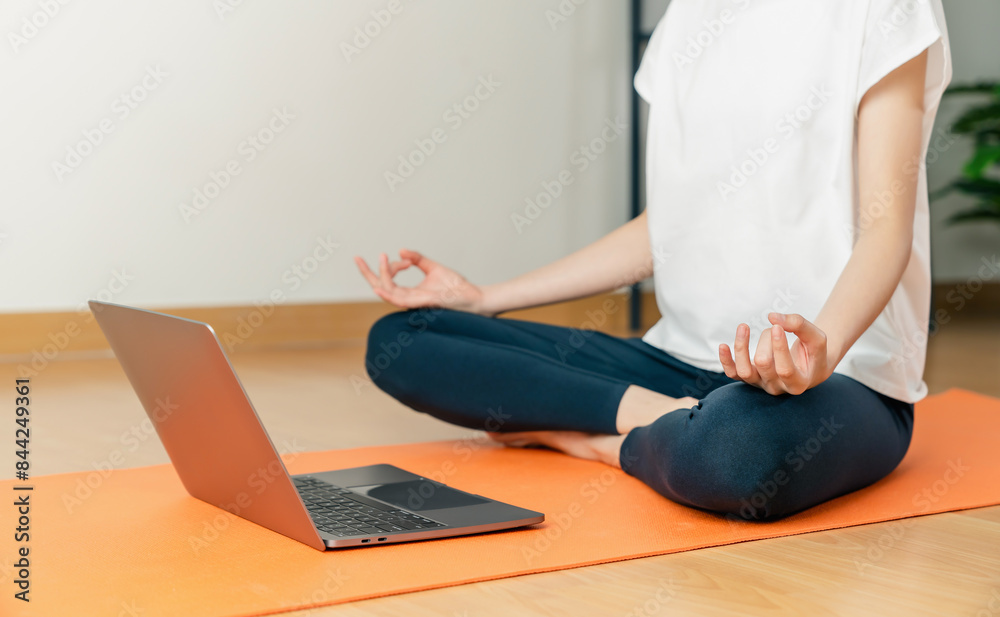 Fototapeta premium Woman sitting on floor in lotus pose and meditating while practicing yoga in home.