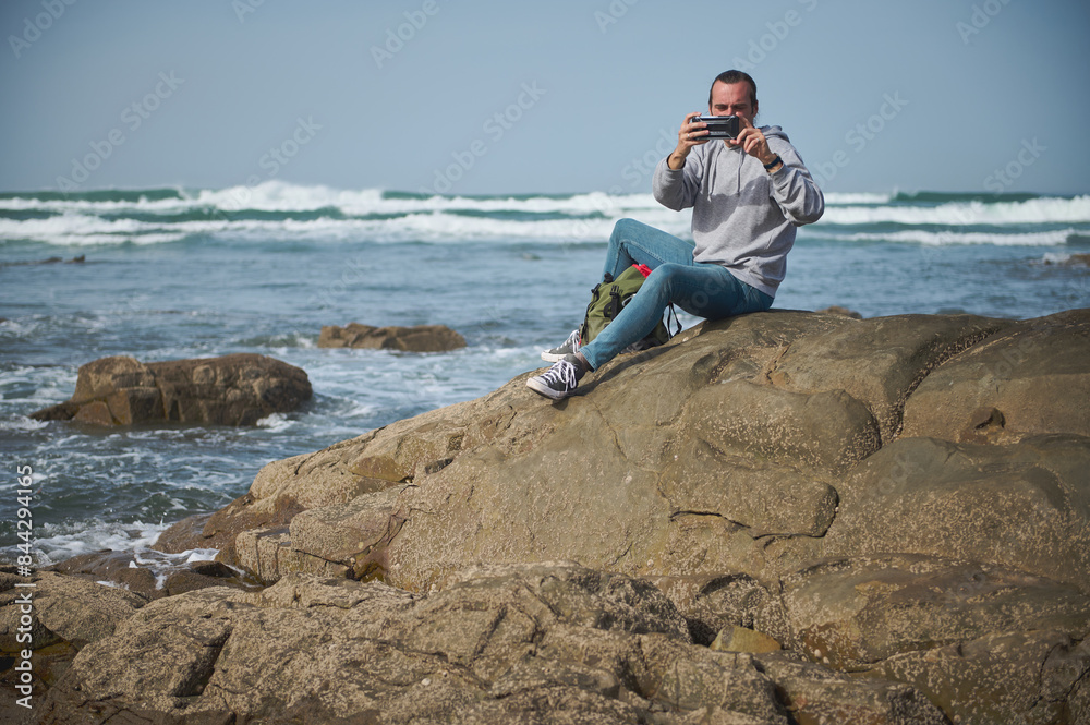 Man taking photos with smartphone on rocky shore by the ocean