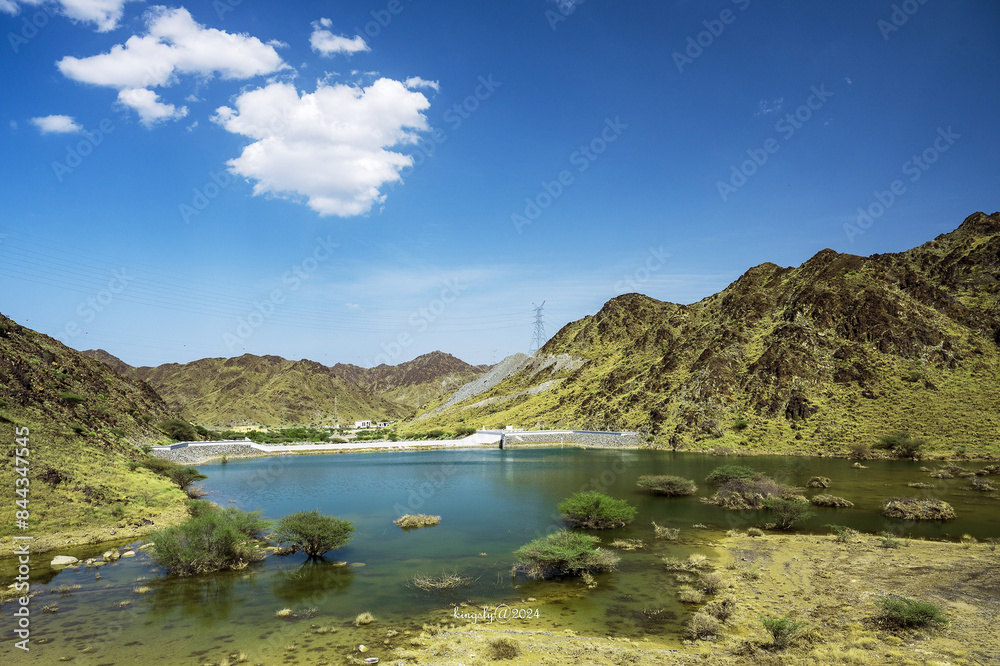 A body of water that is encircled by hills. The outdoor setting features elements like clouds, mountains, and grass. A natural landscape with a water resource is captured in a highland area A body of water that is encircled by hills. The outdoor setting features elements like clouds, mountains, and grass. A natural landscape with a water resource is captured in a highland area