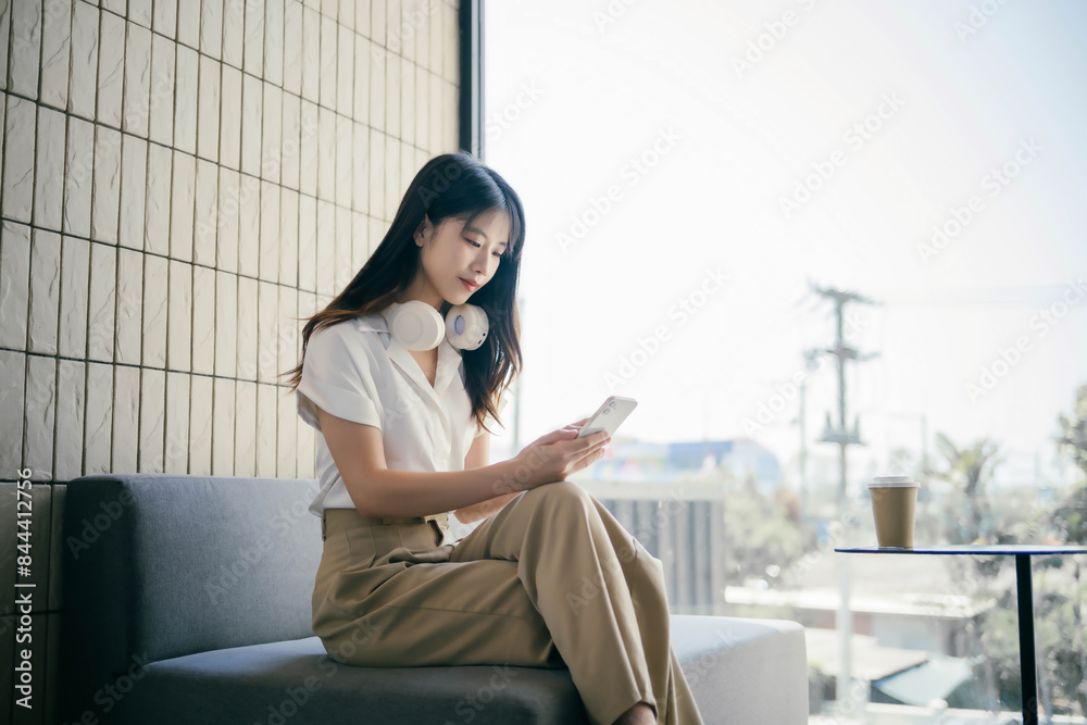 Young Woman Using Smartphone in Modern Office Lounge with Headphones Around Neck, Casual Business Attire, Natural Light, Urban Background, Relaxed Atmosphere, Contemporary Workspace Young Woman Using Smartphone in Modern Office Lounge with Headphones Around Neck, Casual Business Attire, Natural Light, Urban Background, Relaxed Atmosphere, Contemporary Workspace