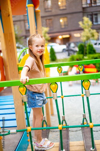 a child girl on a children's multi-colored playground in the courtyard of a multi-storey house plays fun in summer, walks on a cable ladder, summer holidays © Any Grant
